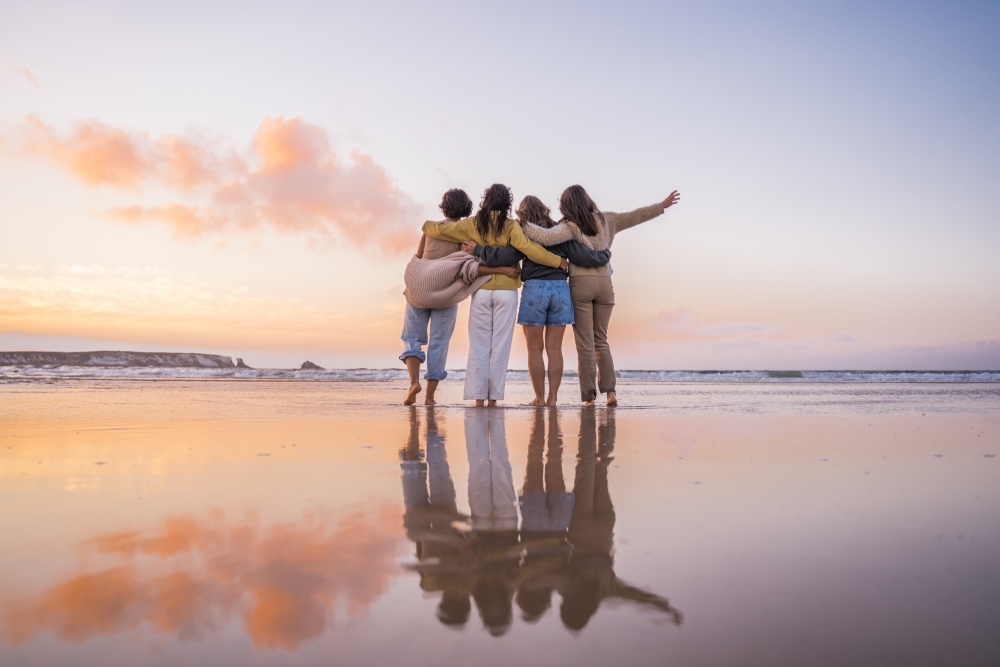 A group of women standing together on the white sands of Jervis Bay at sunrise, silhouetted against a golden and pink sky. They are engaged in conversation and shared presence, representing the biological need for community and co-regulation over digital connection.